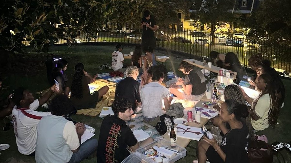 A group of people sitting together on the grass at an outdoor art night, painting and talking