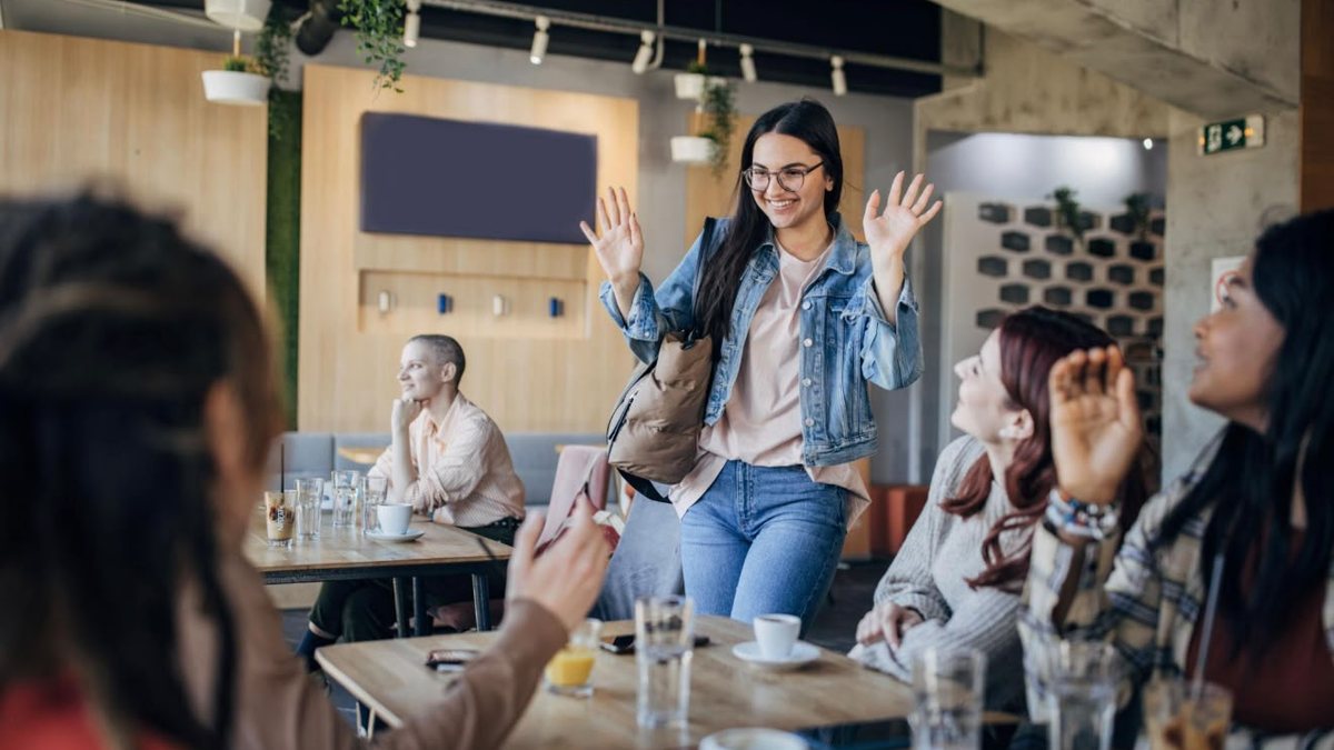 A woman arriving at a cafe and waving hello to the group at her table