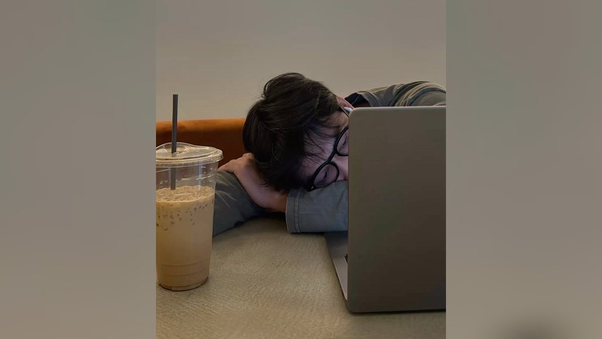 A tired person resting their head on a desk beside a laptop and iced coffee