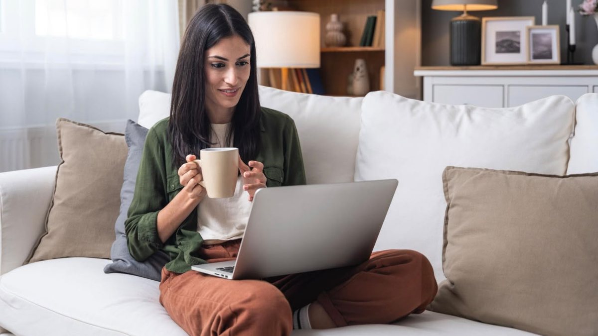 A young woman alone on her sofa with a cup of coffee, looking at her laptop