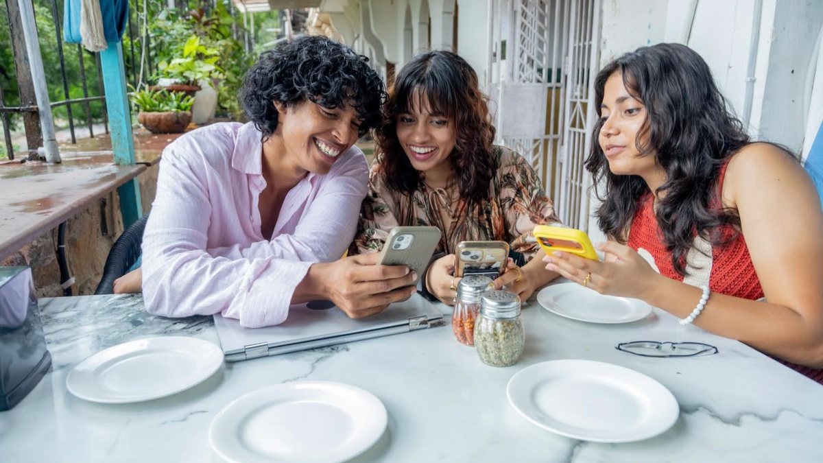 Three friends laughing together at a cafe table while looking at their phones